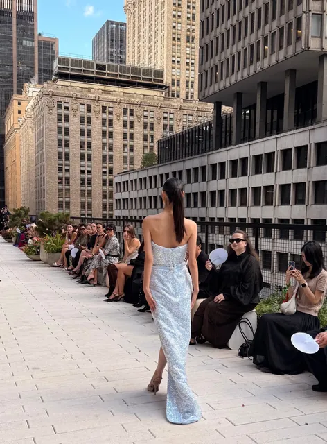 An outdoor fashion runway with a model in a silver gown, seated guests and city buildings.